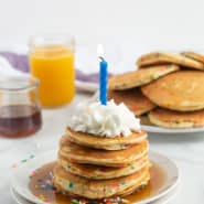 A stack of funfetti pancakes on a plate, topped with syrup, sprinkles, whipped cream, and a blue candle. More pancakes are pictured in the background.
