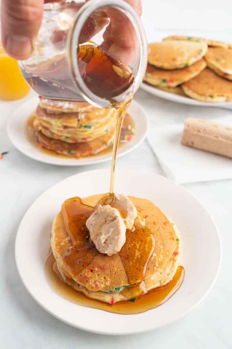 Syrup being poured on pancakes topped with two tabs of flavored butter.