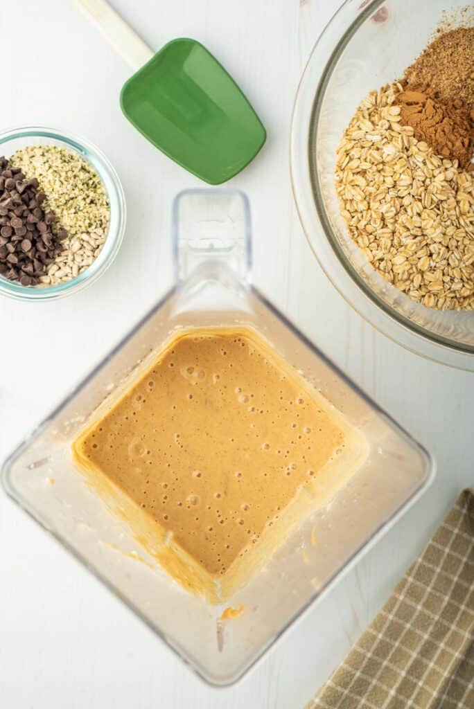 Blender full of light brown substance, surrounded by additional ingredients including oats and chocolate chips.