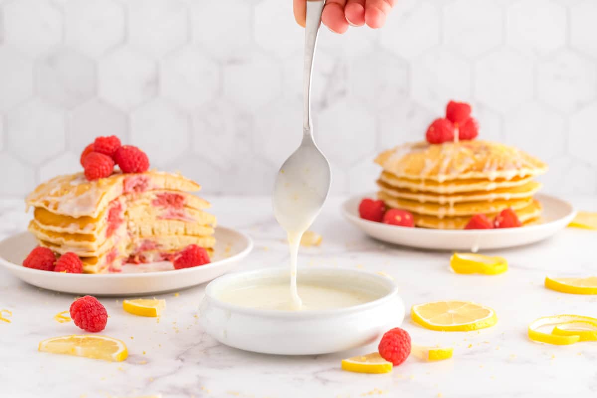 Glaze in a small bowl, pancakes in background. Lemons and raspberries also visible.