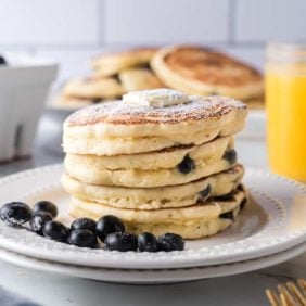 Stack of blueberry pancakes on a white plate, with fresh blueberries. Orange juice in background.