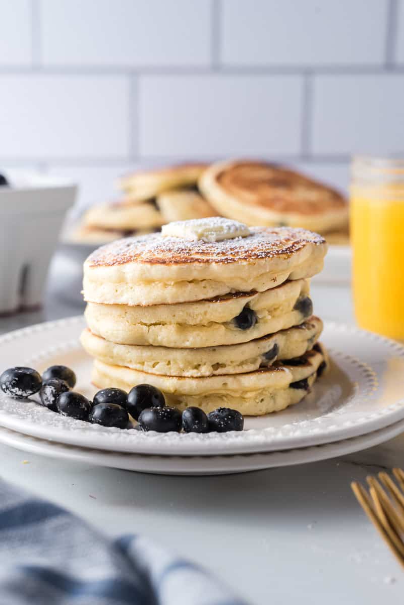 Stack of blueberry pancakes on a white plate, with fresh blueberries. Orange juice in background.