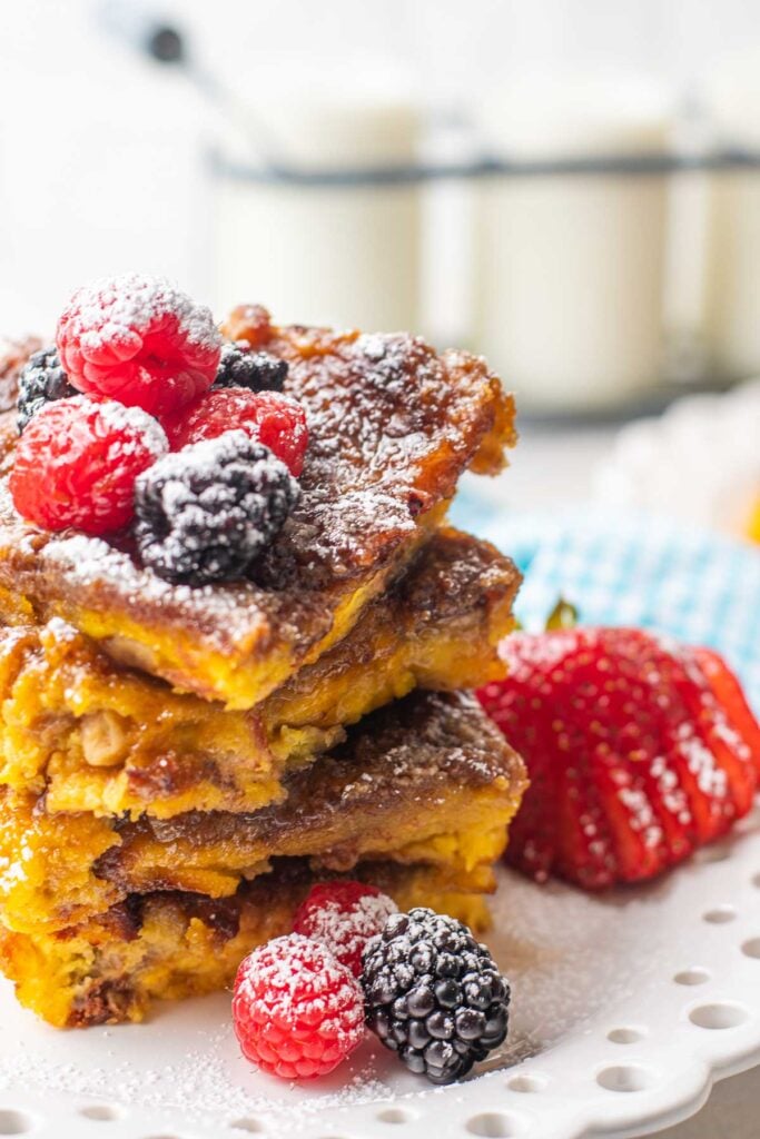 Stack of creme brulee french toast on a white plate with berries and powdered sugar.