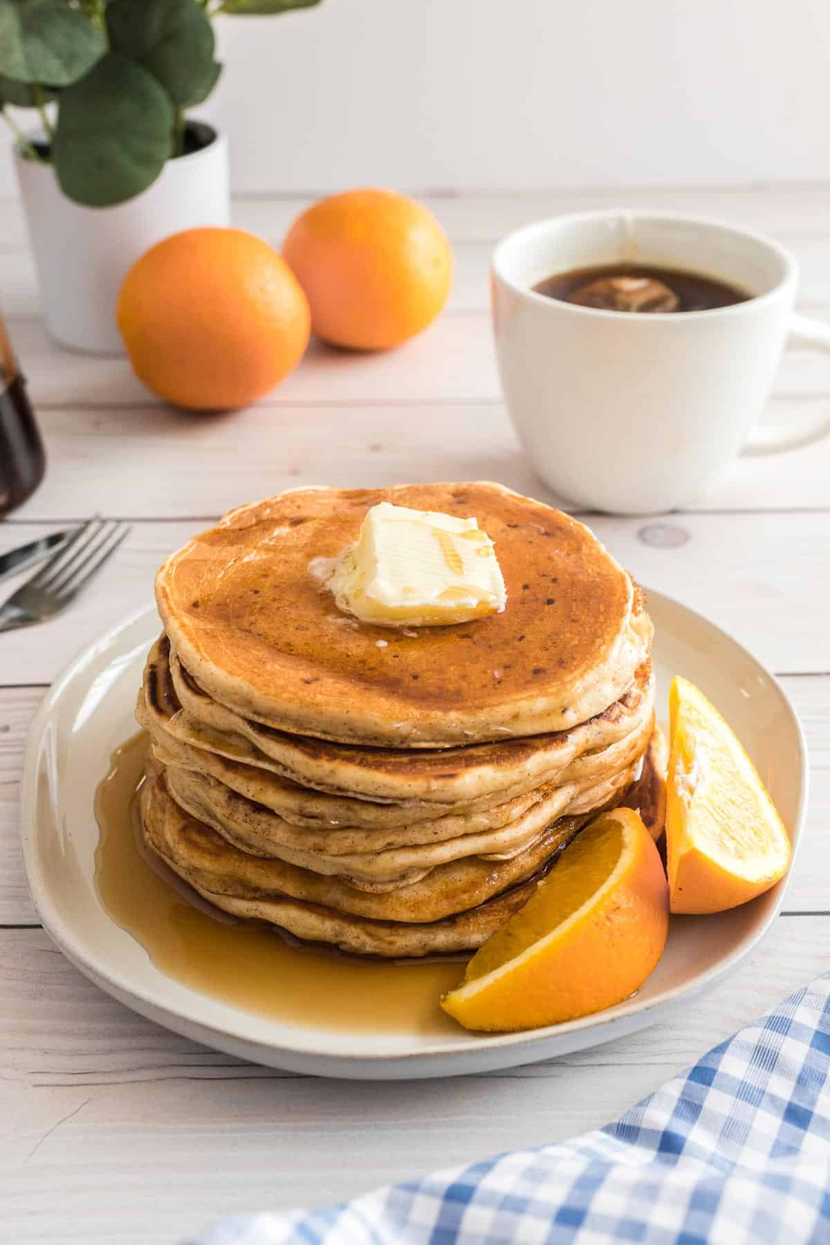 Pancakes and syrup on a plate with orange wedges, coffee and oranges in the background.