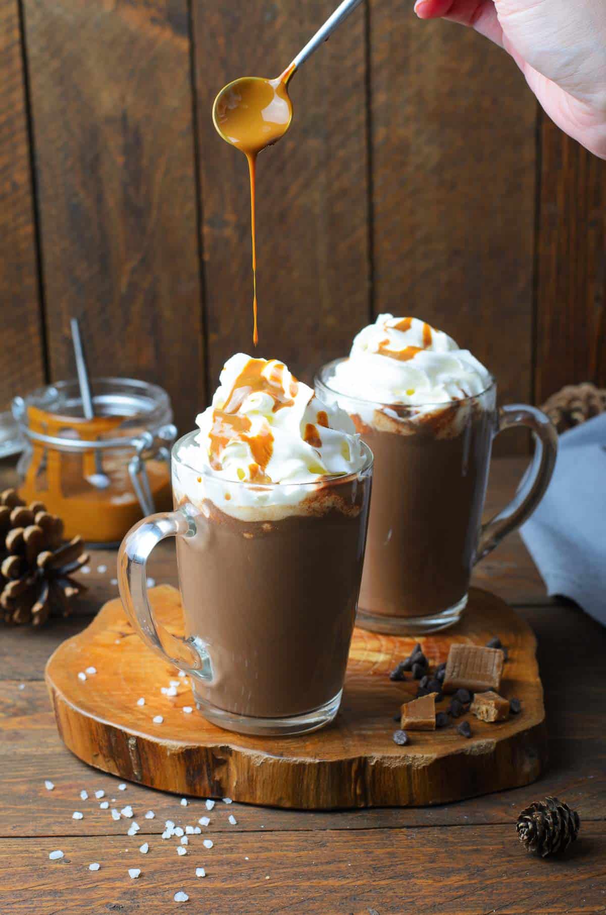 Two clear mugs of hot chocolate topped with whipped cream being drizzled with caramel sauce