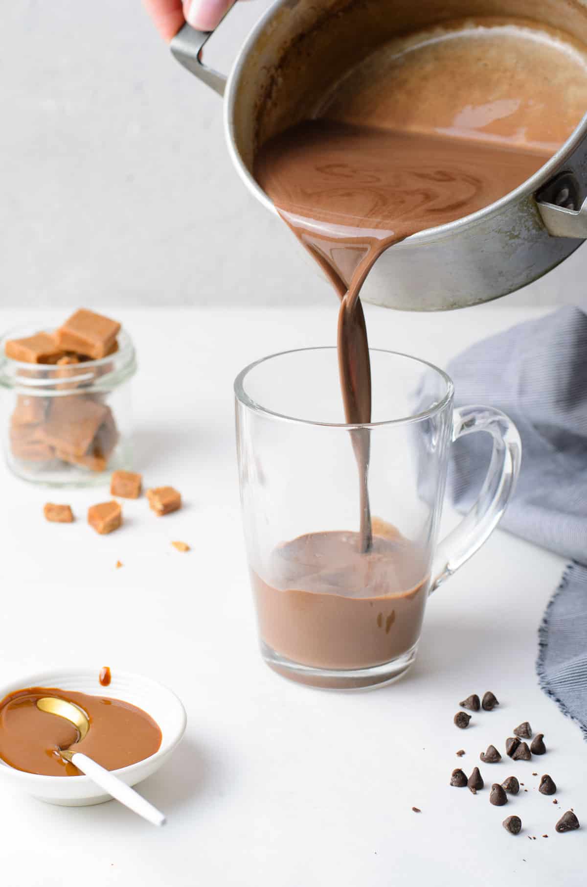 Hot chocolate being poured from a saucepan into a clear glass mug.