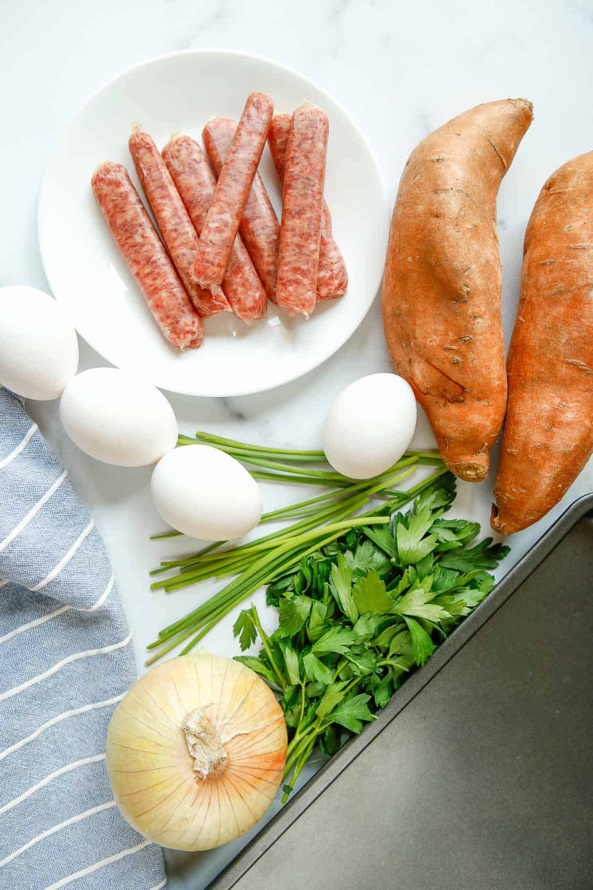 Overhead view of sweet potatoes, sausage, an onion, eggs, and fresh herbs.