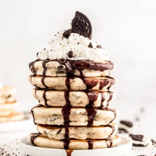 Tall stack of oreo pancakes topped with chocolate syrup, whipped cream, and crushed cookies.