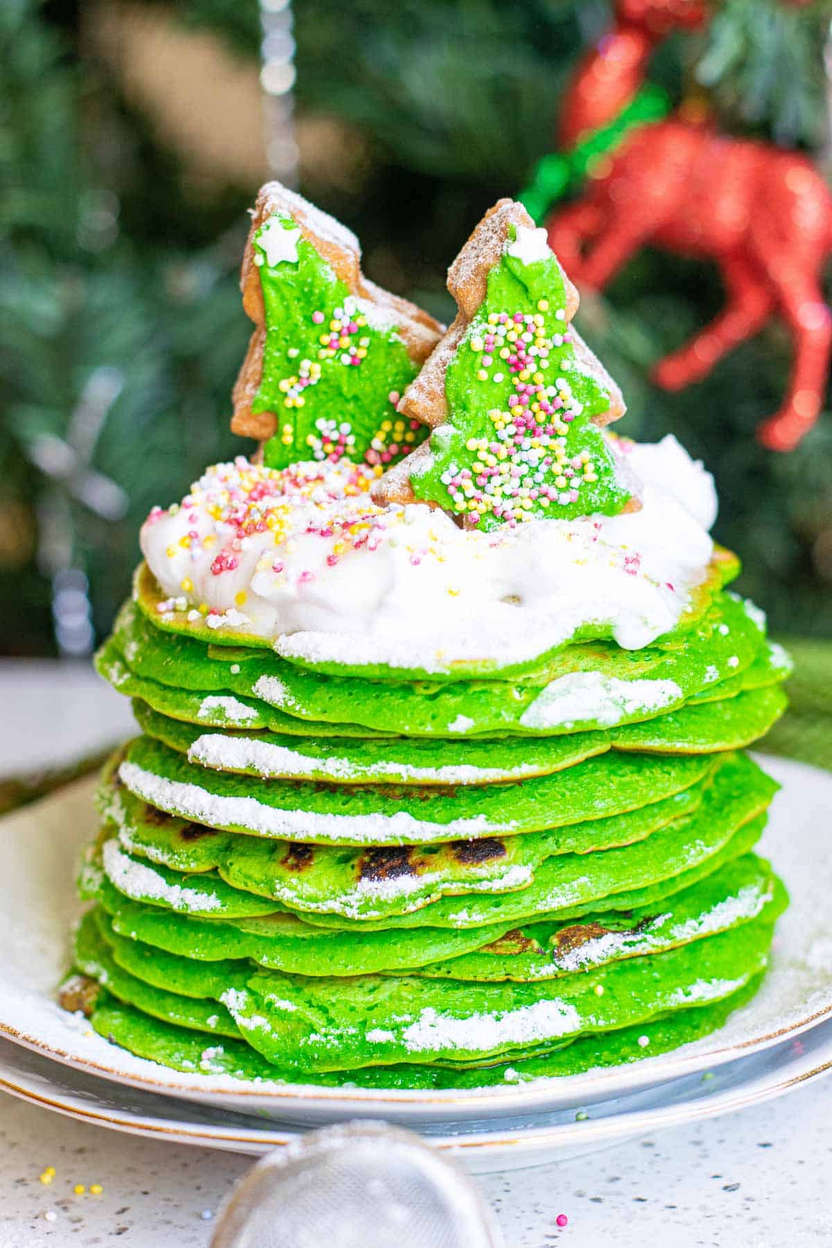 A high stack of bright green pancakes topped with whipped cream and frosted sugar cookies.