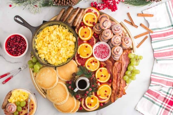 Christmas brunch board with cinnamon rolls, pancakes, scrambled eggs, meat, and fruit.