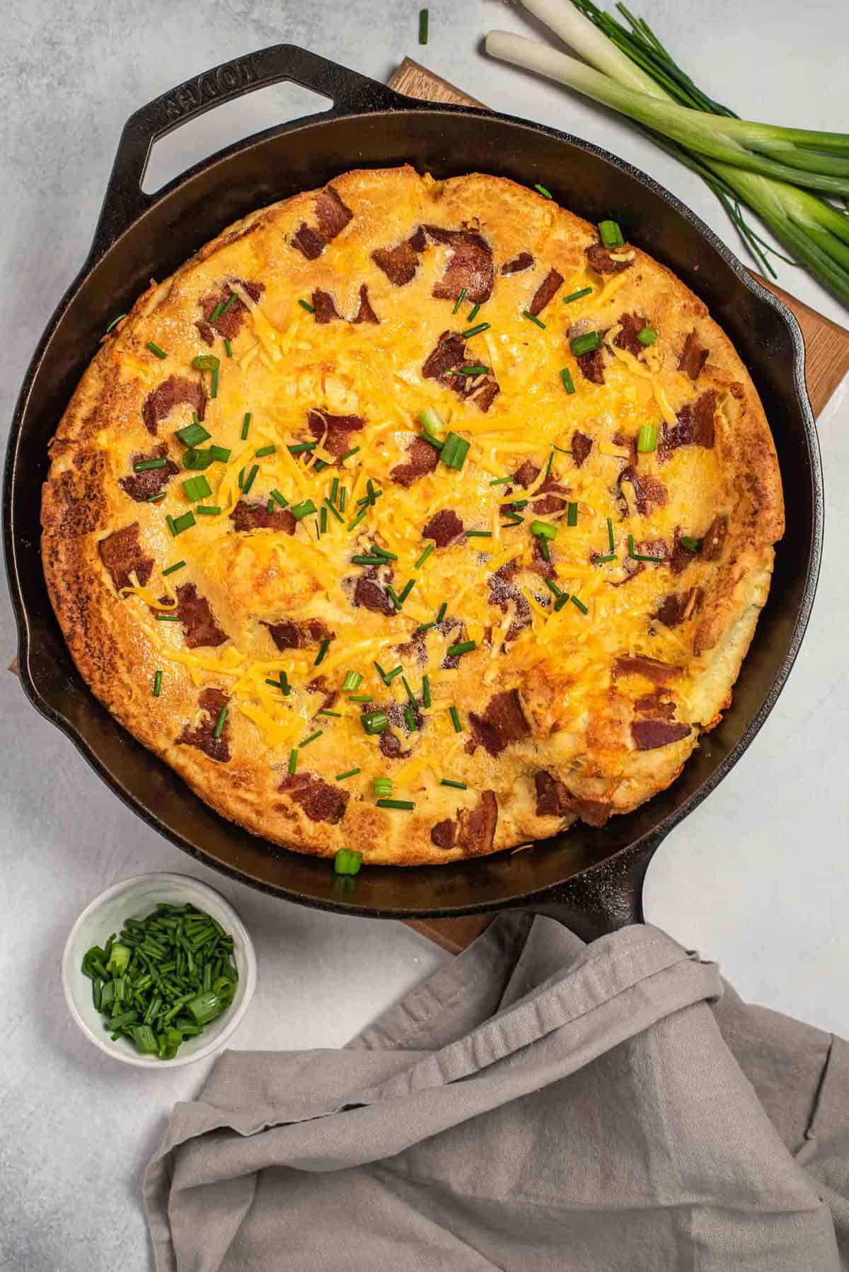 Overhead view of bacon dutch baby in a black skillet garnished with chives.