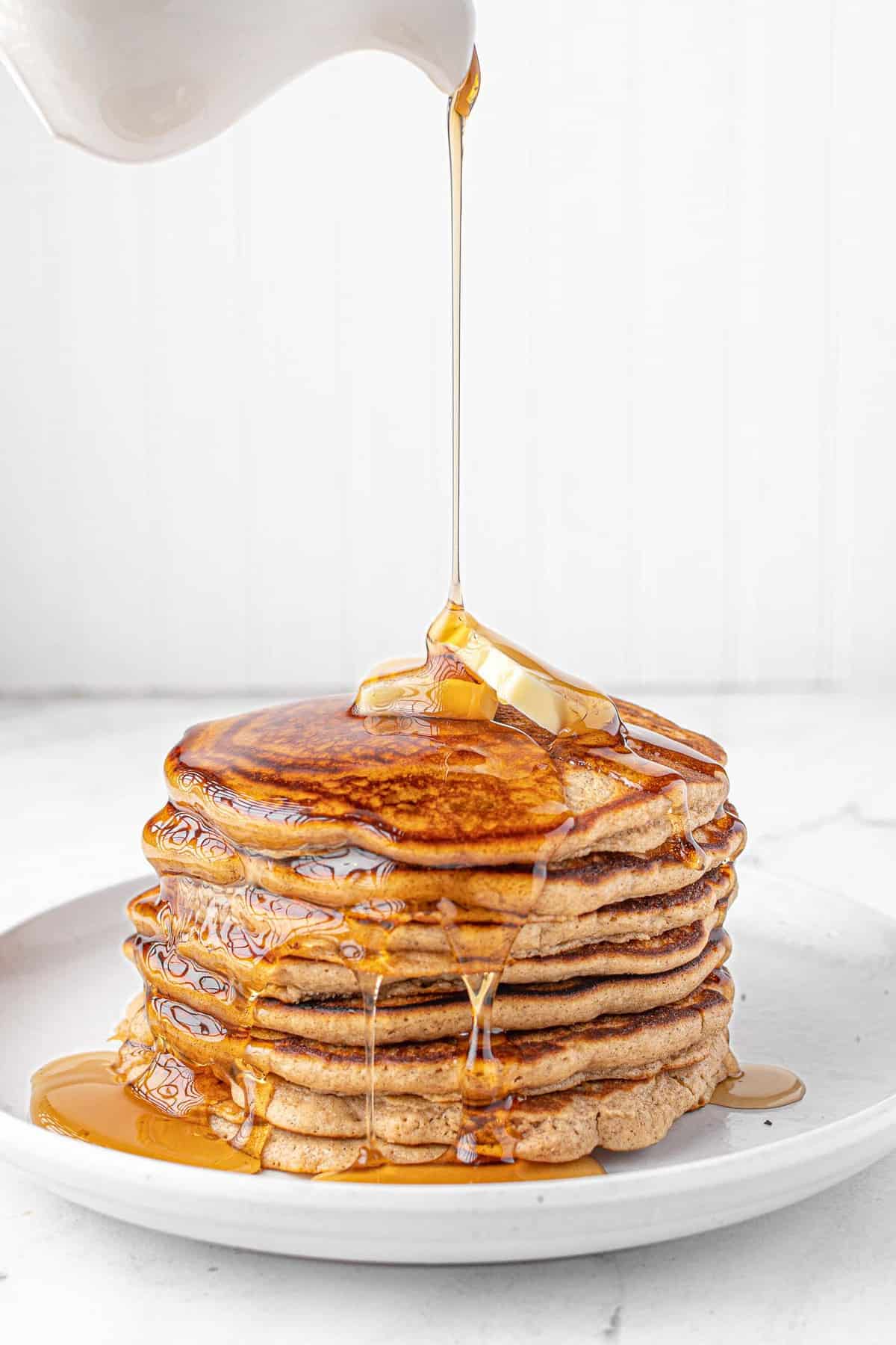 Maple syrup being poured on a large stack of pancakes.