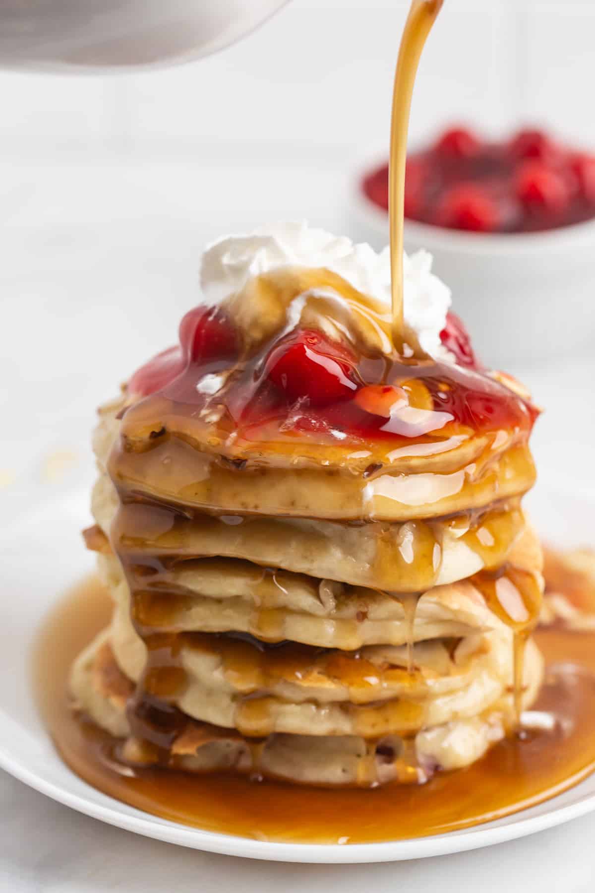 Syrup being poured on a tall stack of pancakes topped with cherry pie filling.