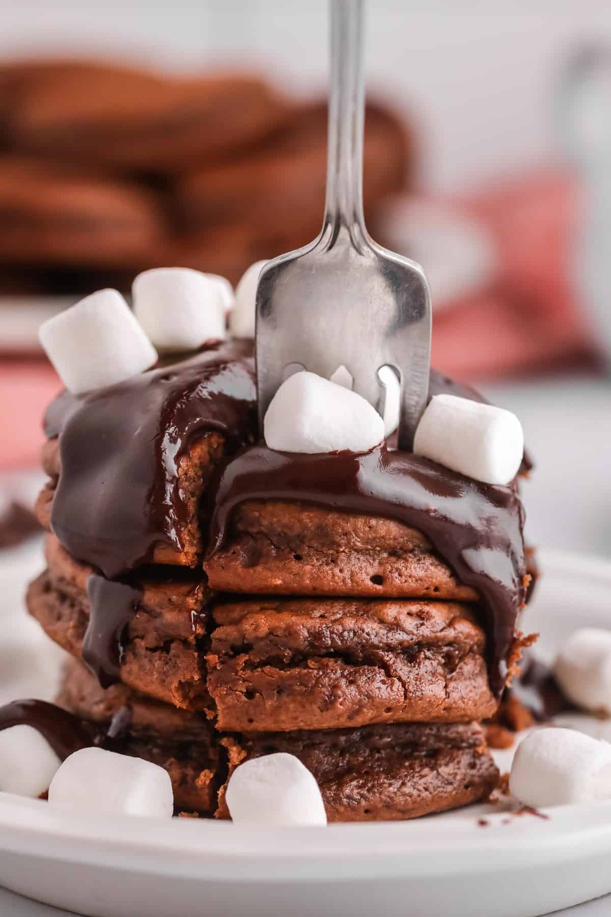 A fork digging into a stack of chocolate pancakes with chocolate syrup.