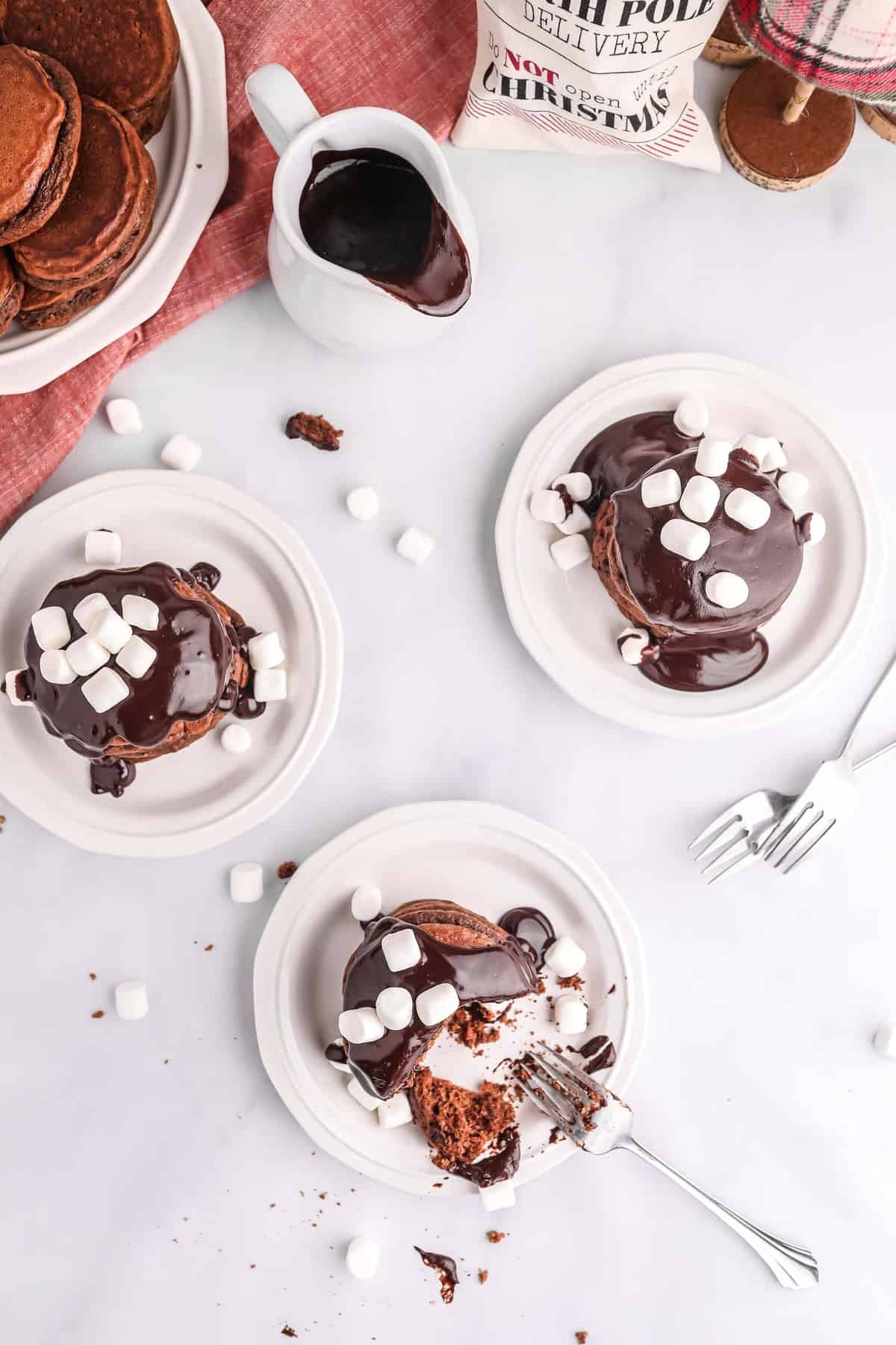 Three plates of pancakes with chocolate sauce and mini marshmallows on top.