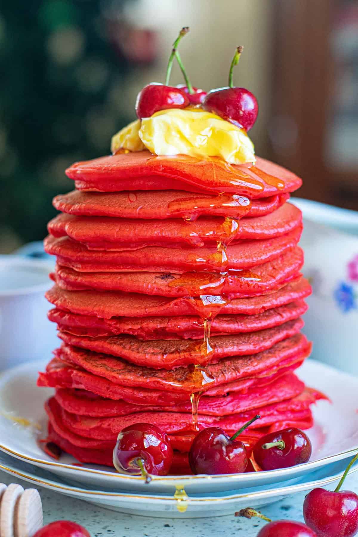 Tall stack of red velvet pancakes topped with cherries and butter.