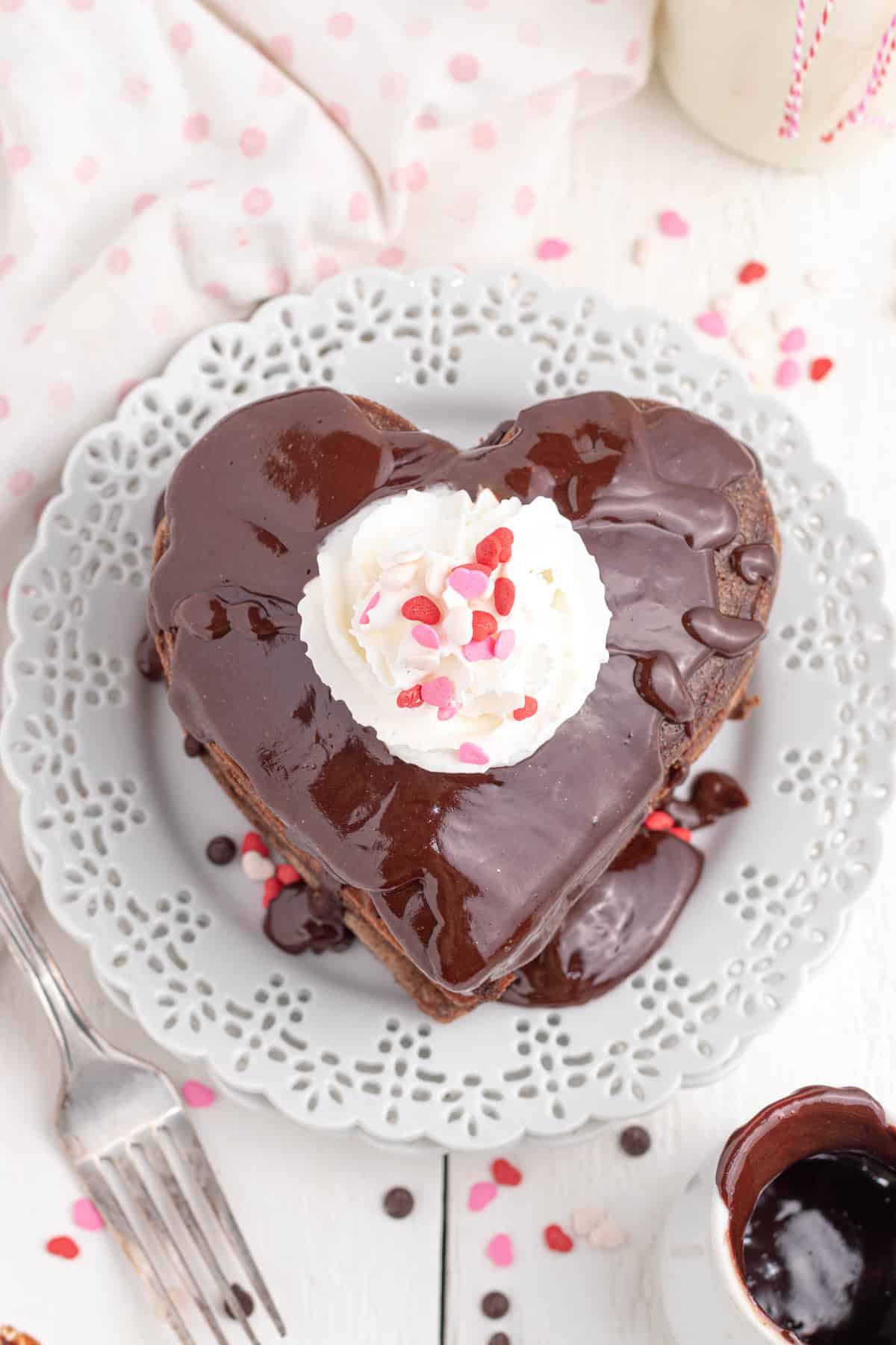 Stack of heart shaped chocolate pancakes topped with ganache, whipped cream, and sprinkles. 