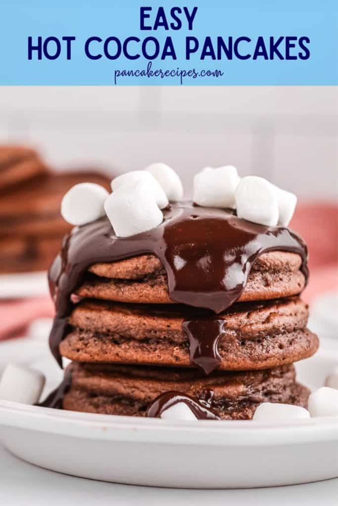 Stack of chocolate pancakes with chocolate sauce and mini marshmallows, text overlay reads "easy hot cocoa pancakes, pancakerecipes.com"