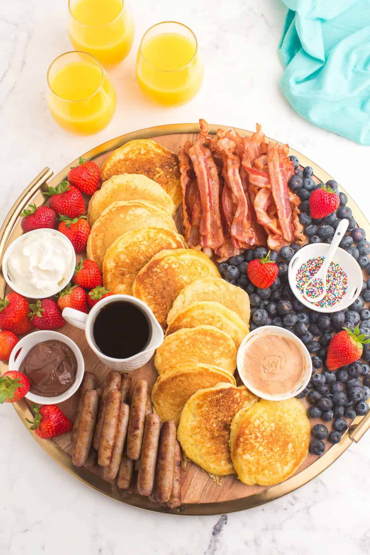 Overhead view of a breakfast spread arranged on a board, including pancakes, bacon, sausage, and fruit.