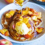 Pancakes, topped with apples and butter, with syrup being poured on top.