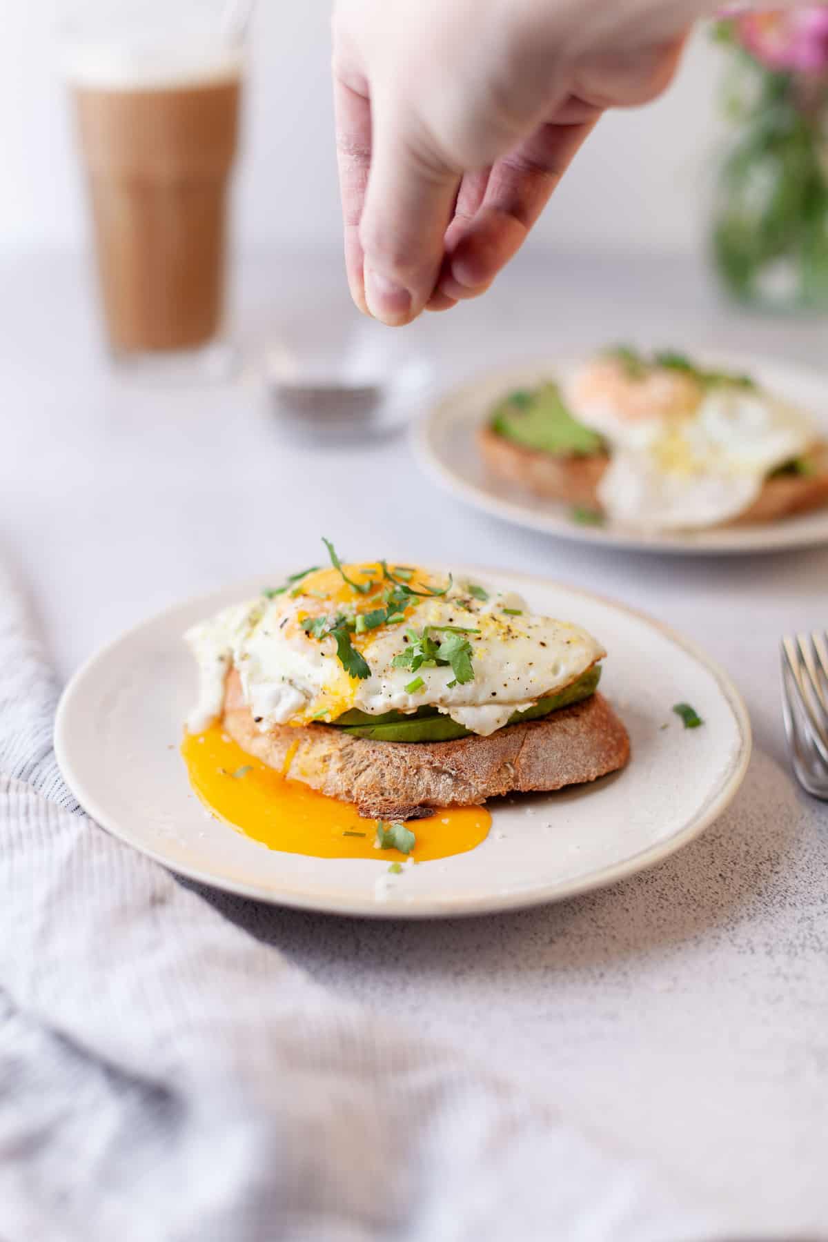 A hand sprinkling herbs on avocado toast with egg.