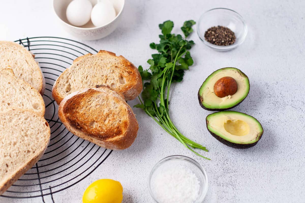 Toast, herbs, avocados, eggs, and seasonings on a grey surface.