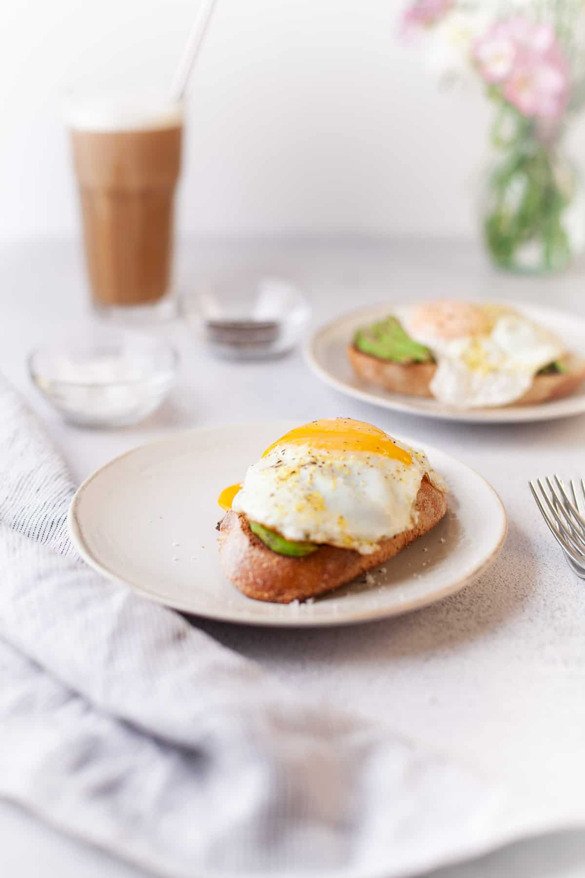 White tabletop with two egg topped avocado toasts, coffee, and a vase of flowers.