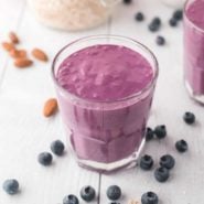 A bright blue smoothie in a short glass, surrounded by fresh blueberries.