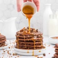 Caramel being poured on a stack of chocolate pancakes with pecans and chocolate chips.