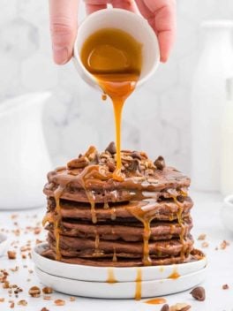 Caramel being poured on a stack of chocolate pancakes with pecans and chocolate chips.