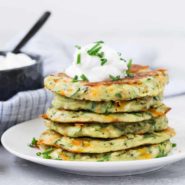 Stack of zucchini fritters on a white plate, topped with sour cream and fresh chives.