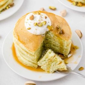 Stack of light green pancakes topped with pistachios and whipped cream.