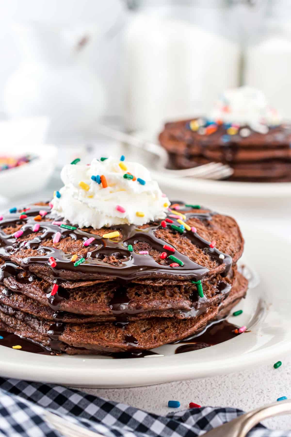 Stack of chocolate pancakes topped with whipped cream, chocolate syrup, and sprinkles.