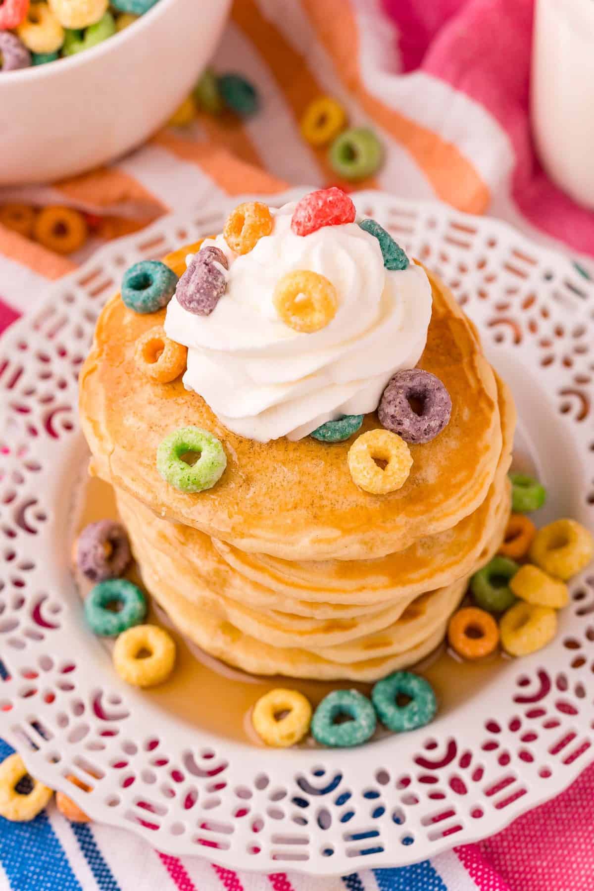 Overhead view of a stack of pancakes topped with froot loops and whipped cream.