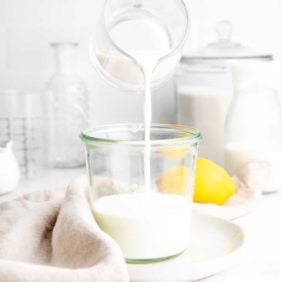 Buttermilk being poured from a small glass pitcher into a small glass jar.
