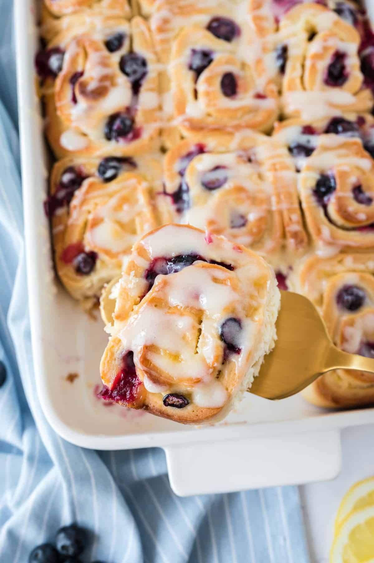 Blueberry roll with glaze being taken out of a pan with a spatula.