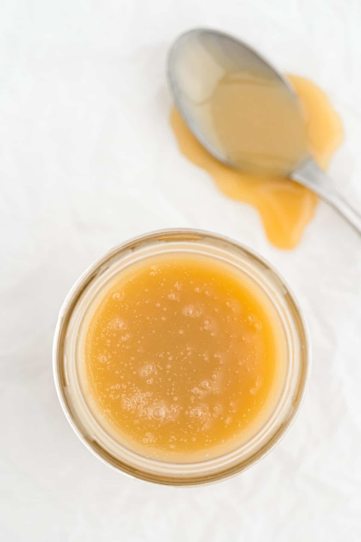 Jar of caramel sauce on a light grey surface, a spoon next to it covered with caramel.