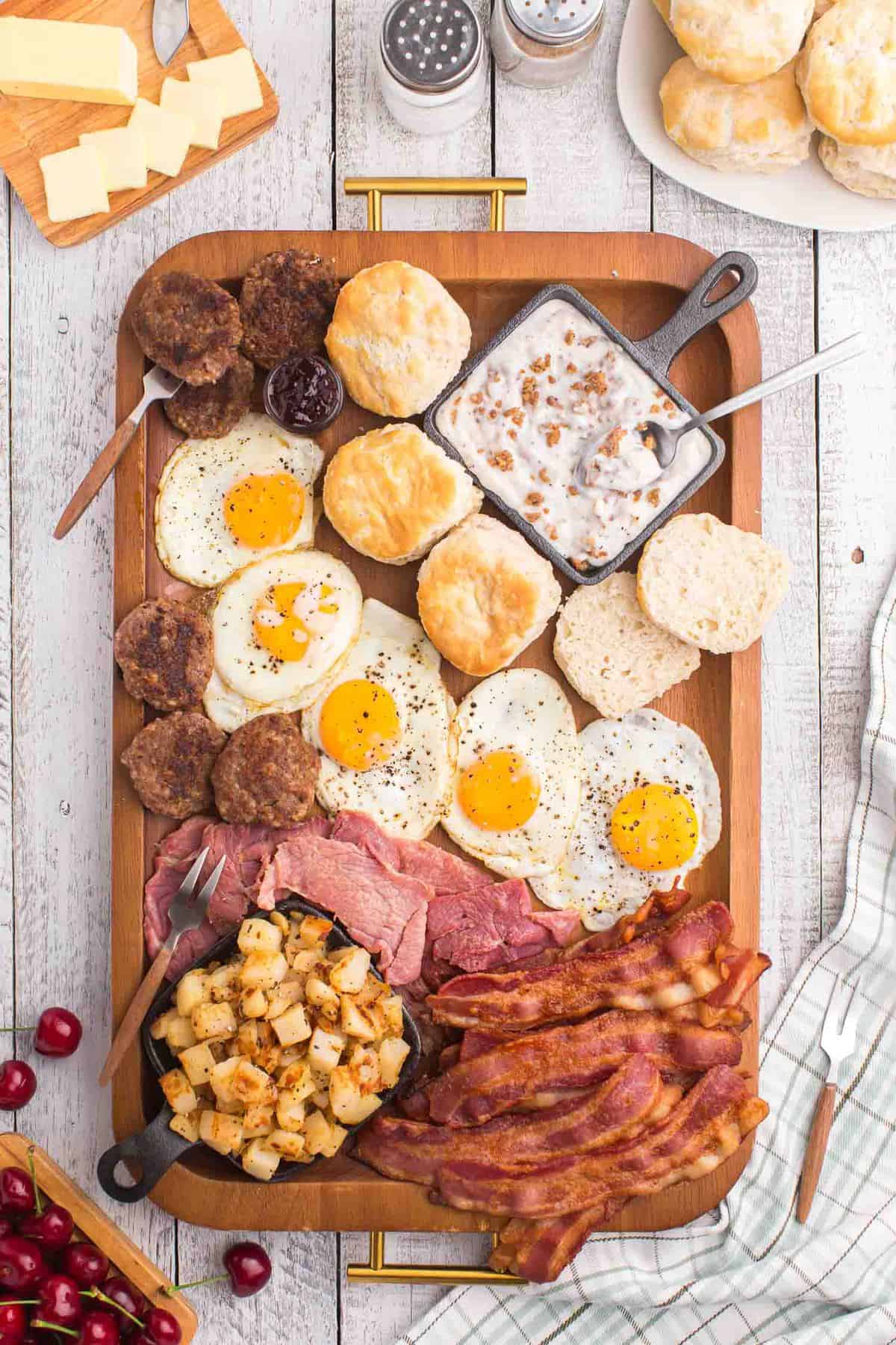Overhead view of meat, eggs, sausage, potatoes, and biscuits on a large wooden tray.