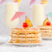 Pineapple pancakes dripping with rum sauce, sprinkled with coconut and topped with a cherry and a paper umbrella.