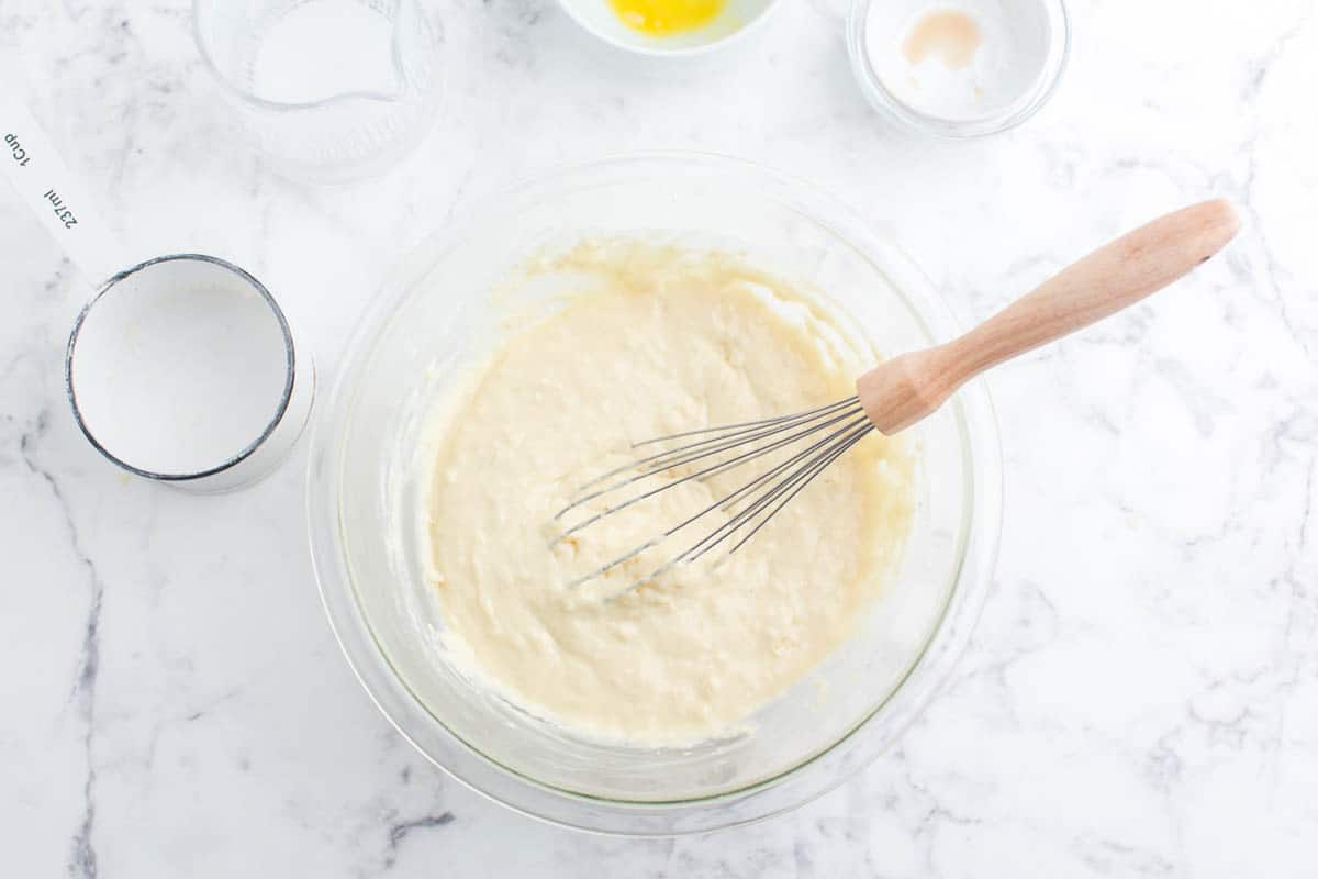 Pancake batter in a clear bowl with a wooden handled whisk.