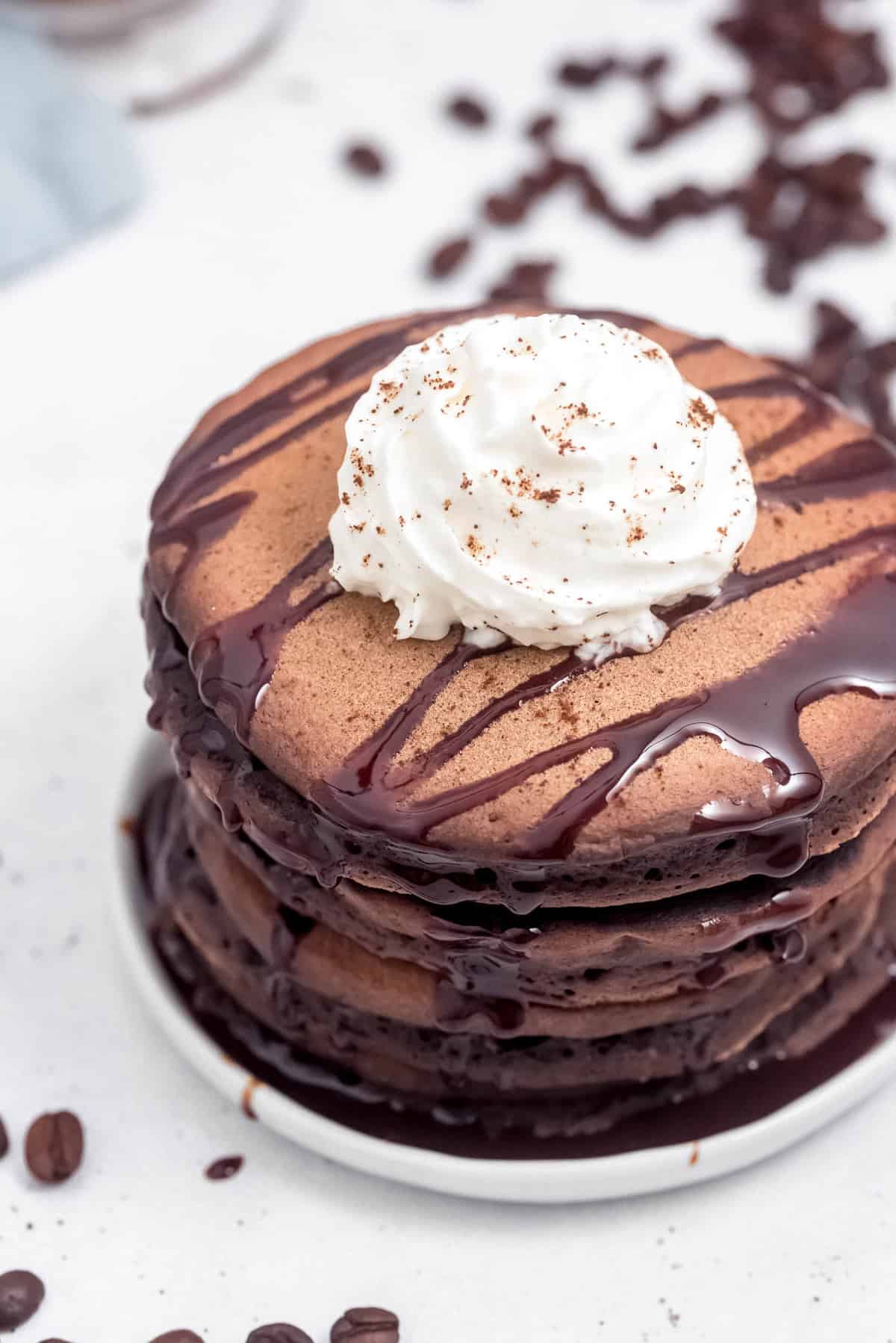 Stack of dark brown chocolate pancakes topped with whipped cream and chocolate syrup, on a white plate.