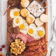 Overhead view of meat, eggs, sausage, potatoes, and biscuits on a large wooden tray.