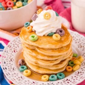 Tall stack of pancakes topped with whipped cream and froot loops cereal.