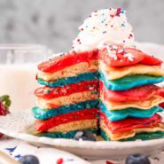 Red, white, and blue pancakes on a white plate, topped with whipped cream and sprinkles.