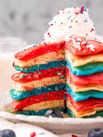 Red, white, and blue pancakes on a white plate, topped with whipped cream and sprinkles.