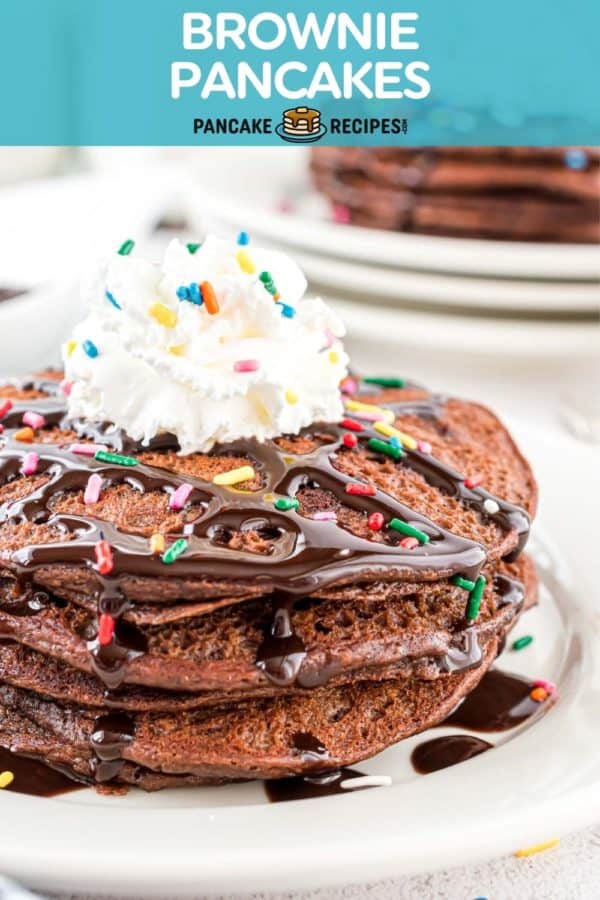 Stack of chocolate pancakes topped with whipped cream, chocolate syrup, and sprinkles. Text overlay reads "brownie pancakes, pancakerecipes.com."