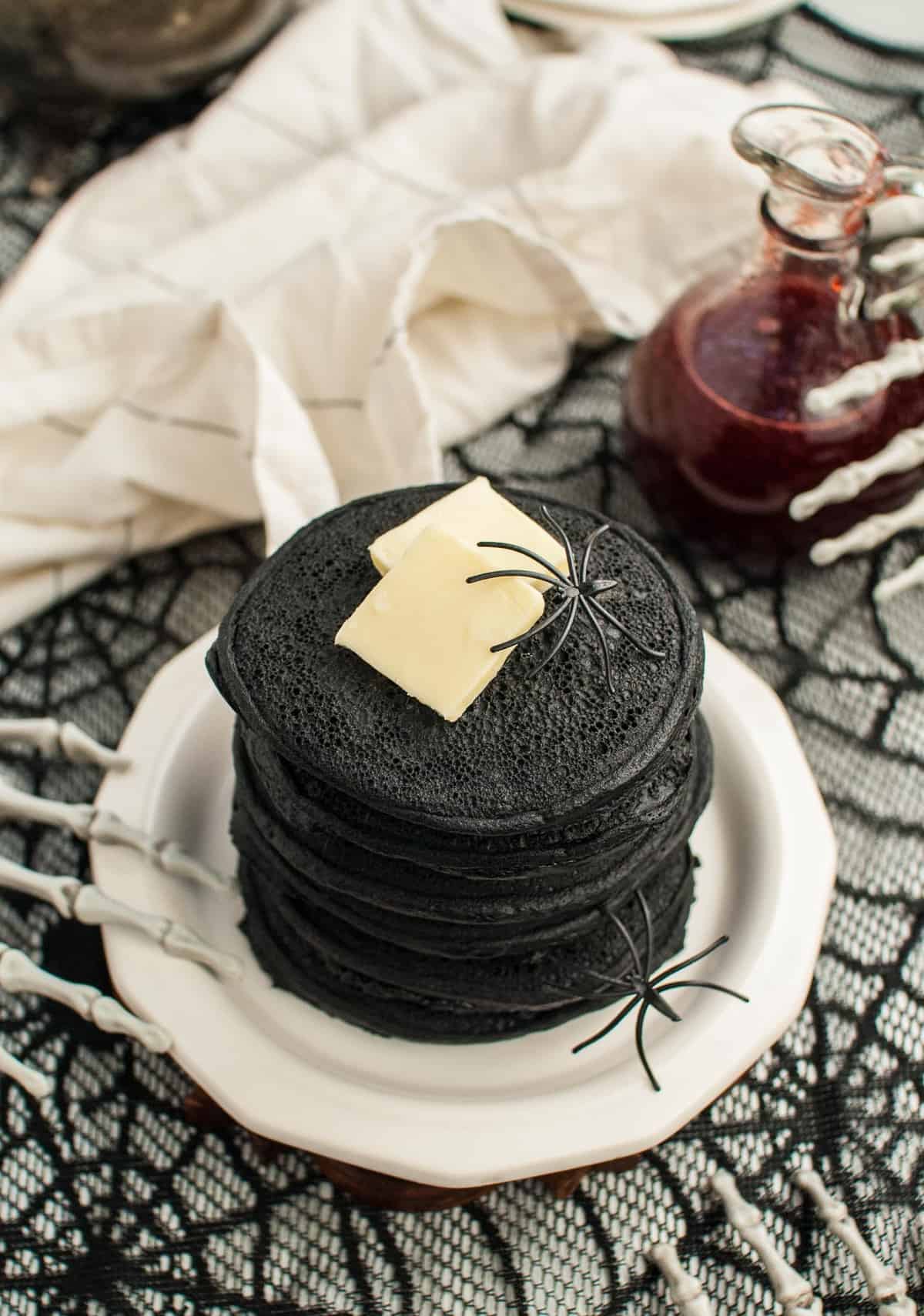 Dark black pancakes in a stack, topped with butter, surrounded by skeleton hands and fake spiders.