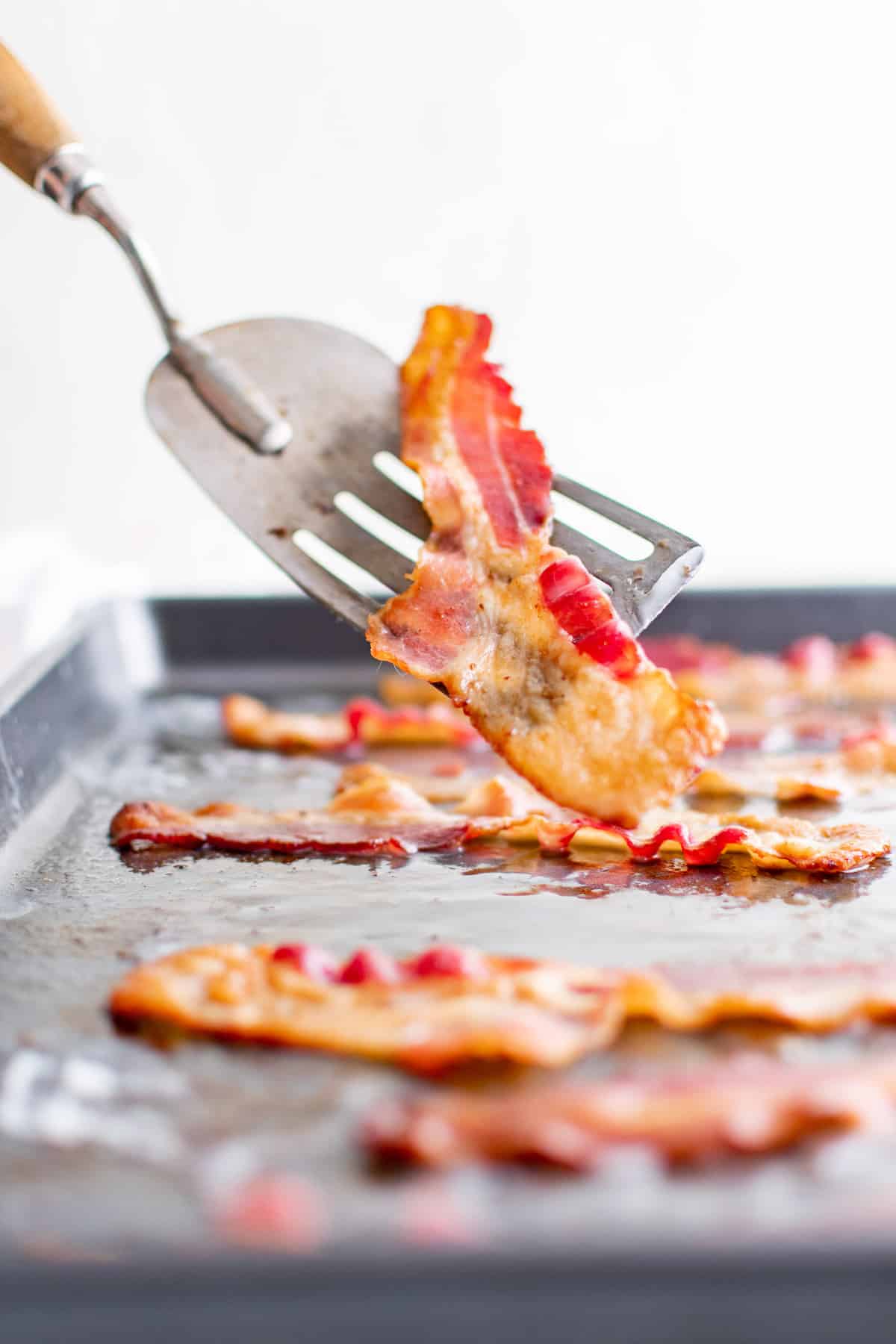 Crispy bacon on a spatula, more on a sheet pan in the background.