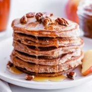 Stack of apple butter pancakes dripping with syrup and topped with pecans.
