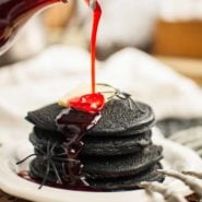 Stack of dark black pancakes topped with butter, a red syrup being poured on top.